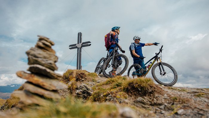 Zwei Fahrradfahrer mit Helmen auf einem Berggipfel neben einem Kreuz