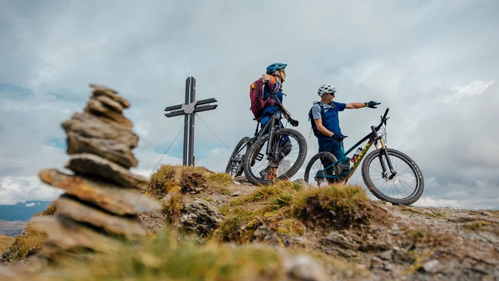 Spätsommer- & Herbstbiken in Toblach © Felix Saller Zwei Fahrradfahrer mit Helmen auf einem Berggipfel neben einem Kreuz
