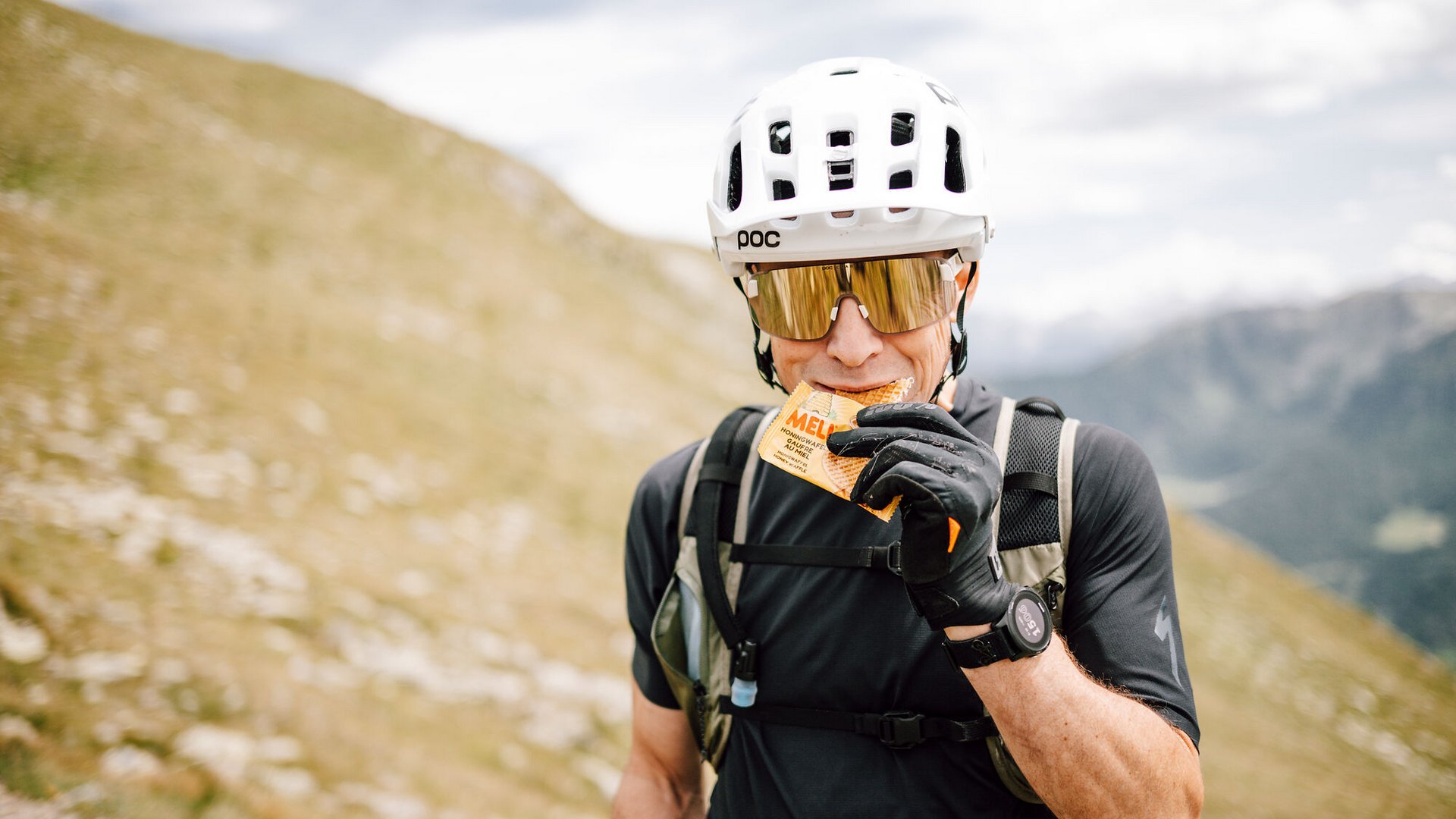 Mann mit Fahrradhelm isst Energieriegel in bergiger Landschaft