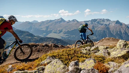 Zwei Mountainbiker fahren auf einem Bergpfad mit Alpen im Hintergrund
