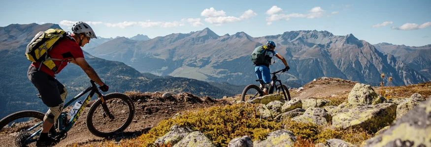 Zwei Mountainbiker fahren auf einem Bergpfad mit Alpen im Hintergrund