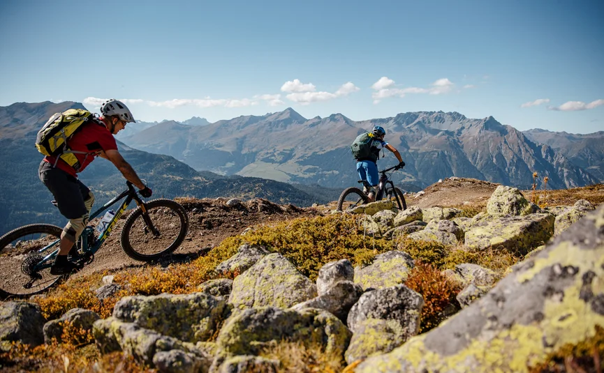 Two mountain bikers riding on a mountain trail with alpine mountains behind