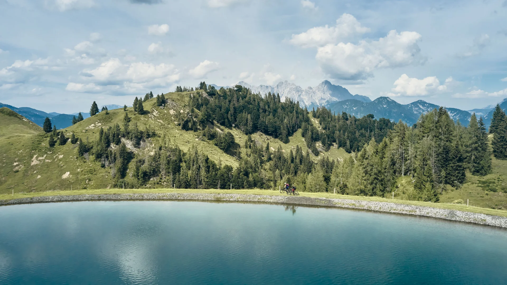 Ready to ride: Das ist neu in Österreichs größter Bike-Region © Tobias Köhler Zwei Radfahrer entlang eines Bergsees mit bewaldeten Hügeln und Bergen im Hintergrund