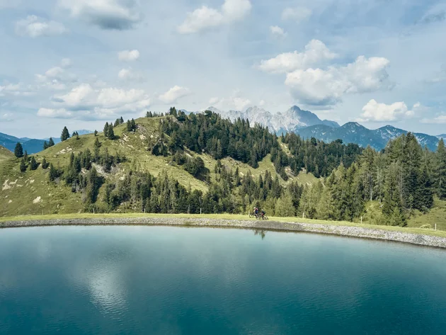 Happy Birthday EPIC Bike Park! © Tobias Köhler Zwei Radfahrer entlang eines Bergsees mit bewaldeten Hügeln und Bergen im Hintergrund