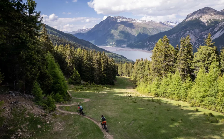 Zwei Mountainbiker fahren auf einem Pfad durch bewaldete Berge mit See im Hintergrund