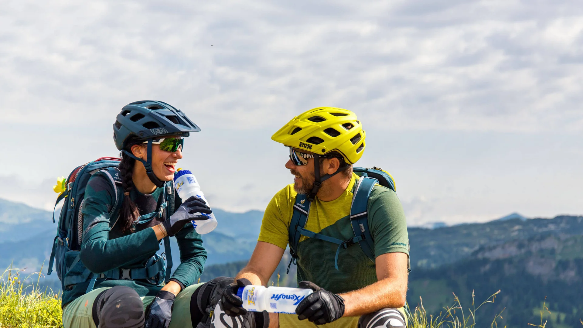 Zwei Radfahrer pausieren lachend auf einer Bergwiese mit Trinkflaschen