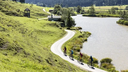 Vier Radfahrer auf einem Weg entlang eines Sees in hügeliger Landschaft