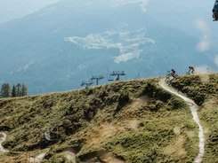 Zwei Fahrradfahrer auf einem Bergpfad mit Sessellift vor bewaldeten Bergen