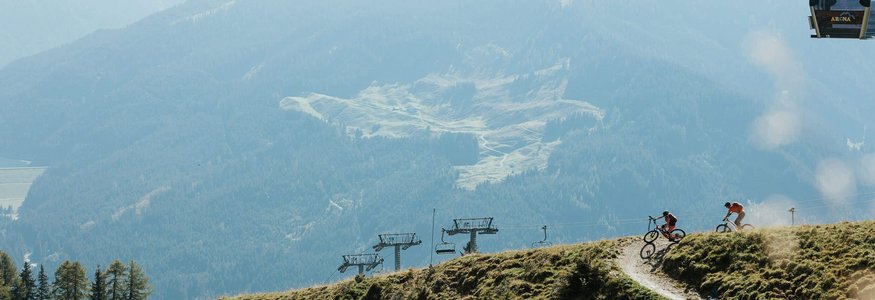 Zwei Fahrradfahrer auf einem Bergpfad mit Sessellift vor bewaldeten Bergen