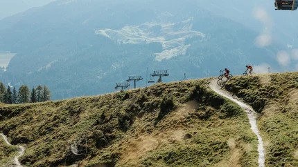 Twee mountainbikers op een bergpad met stoellift en beboste bergen op de achtergrond