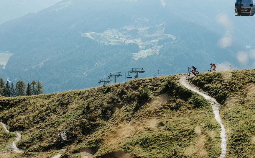 Zwei Fahrradfahrer auf einem Bergpfad mit Sessellift vor bewaldeten Bergen