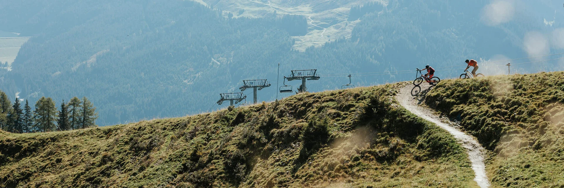Twee mountainbikers op een bergpad met stoellift en beboste bergen op de achtergrond
