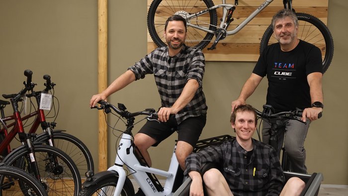 Jubiläumsjahr 20/25 Jahre „Bikestore.cc“ © Bikestore.cc Drei Männer mit Fahrrädern, einer sitzt in einem Lastenradkorb