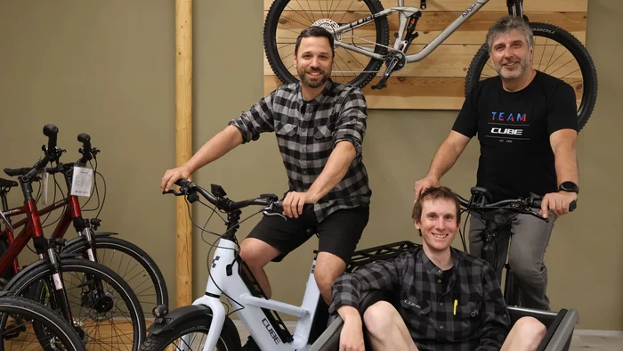 Drei Männer mit Fahrrädern, einer sitzt in einem Lastenradkorb