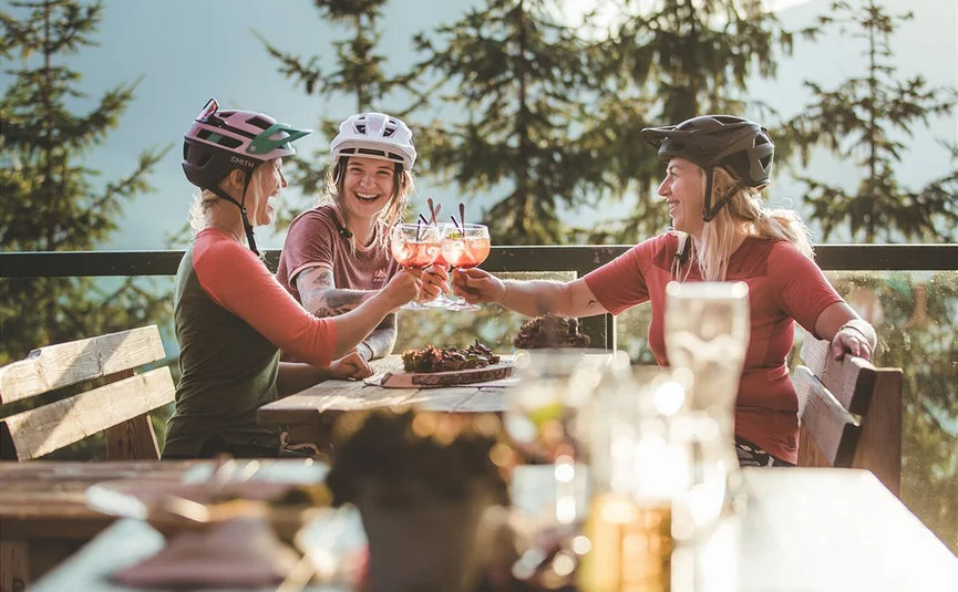 Three cyclists toast with drinks on a forest terrace in sunlight