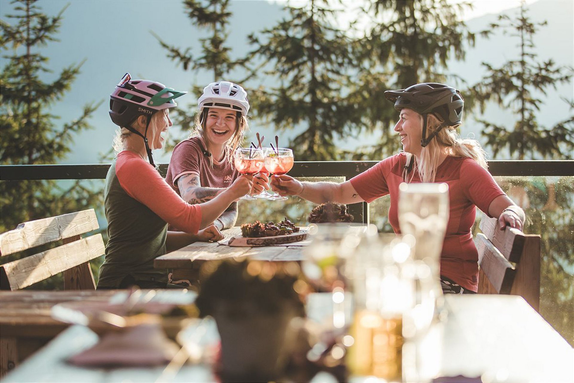 Drei Radfahrerinnen stoßen mit Getränken an auf einer Terrasse im Wald