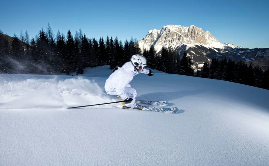 4 Tage Genuss-Skifahren auf der Sonnenseite der Zugspitze 2026-7 Skifahrer in weißer Kleidung fährt auf frischem Schnee mit Berg und Wald im Hintergrund