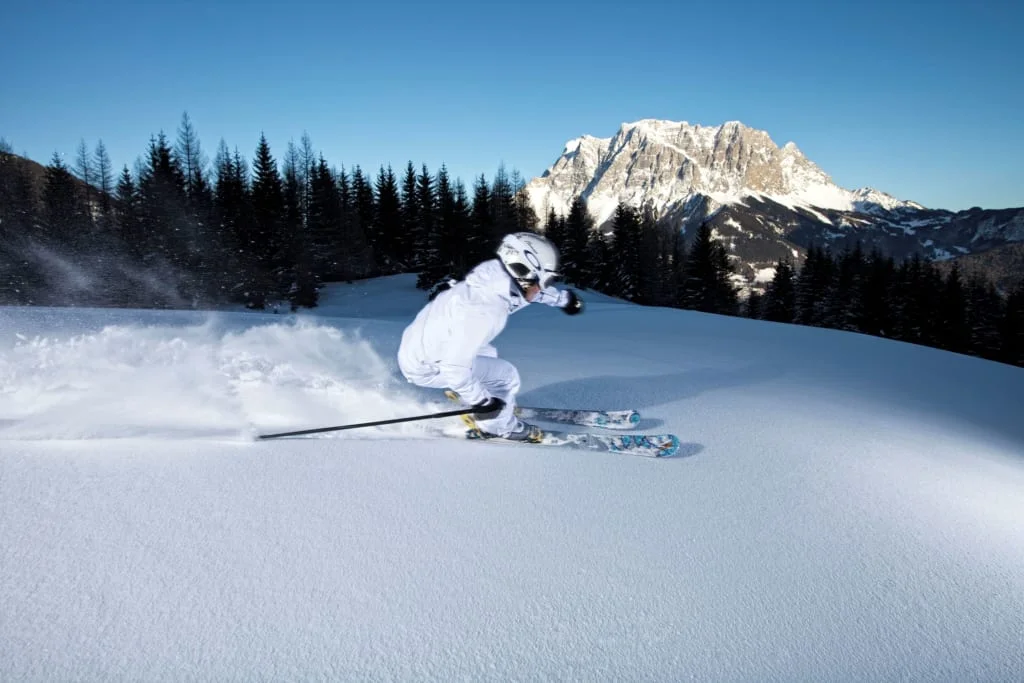 Skier in white outfit skiing on fresh snow with mountain and forest backdrop
