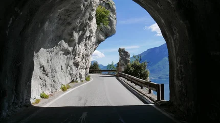 Bergstraße mit Tunnelöffnung und Blick auf See und Berge