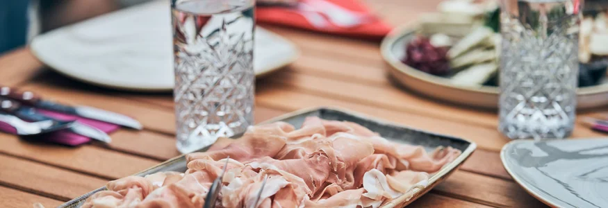 Auf einem Holztisch liegt ein Teller mit dünn geschnittenem Schinken und Besteck