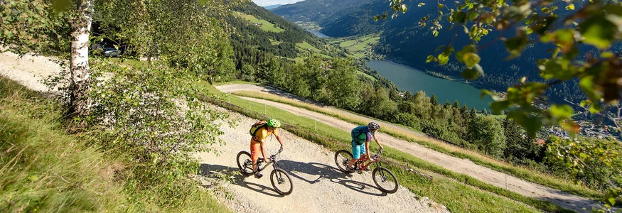 Zwei Fahrradfahrer auf einem Bergweg mit Blick auf Tal und See