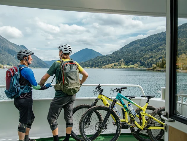 Mountainbiken im Herbst: 5 Top-Regionen für die Saisonverlängerung © David Karg Zwei Radfahrer auf einer Fähre mit Bergsee und Bergen im Hintergrund