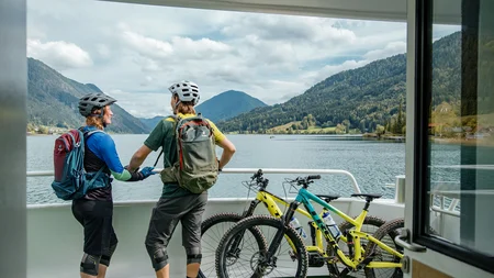 Mountainbiken im Herbst: 5 Top-Regionen für die Saisonverlängerung © David Karg Zwei Radfahrer auf einer Fähre mit Bergsee und Bergen im Hintergrund