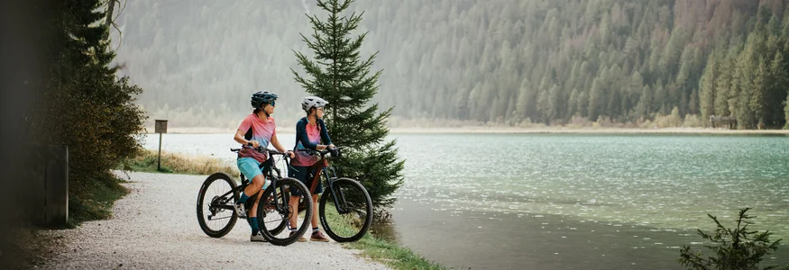 Spätsommer- & Herbstbiken in Toblach © Desire Lines - Tobias Dörr Zwei Radfahrer auf einem Waldweg neben einem See mit Bergen im Hintergrund