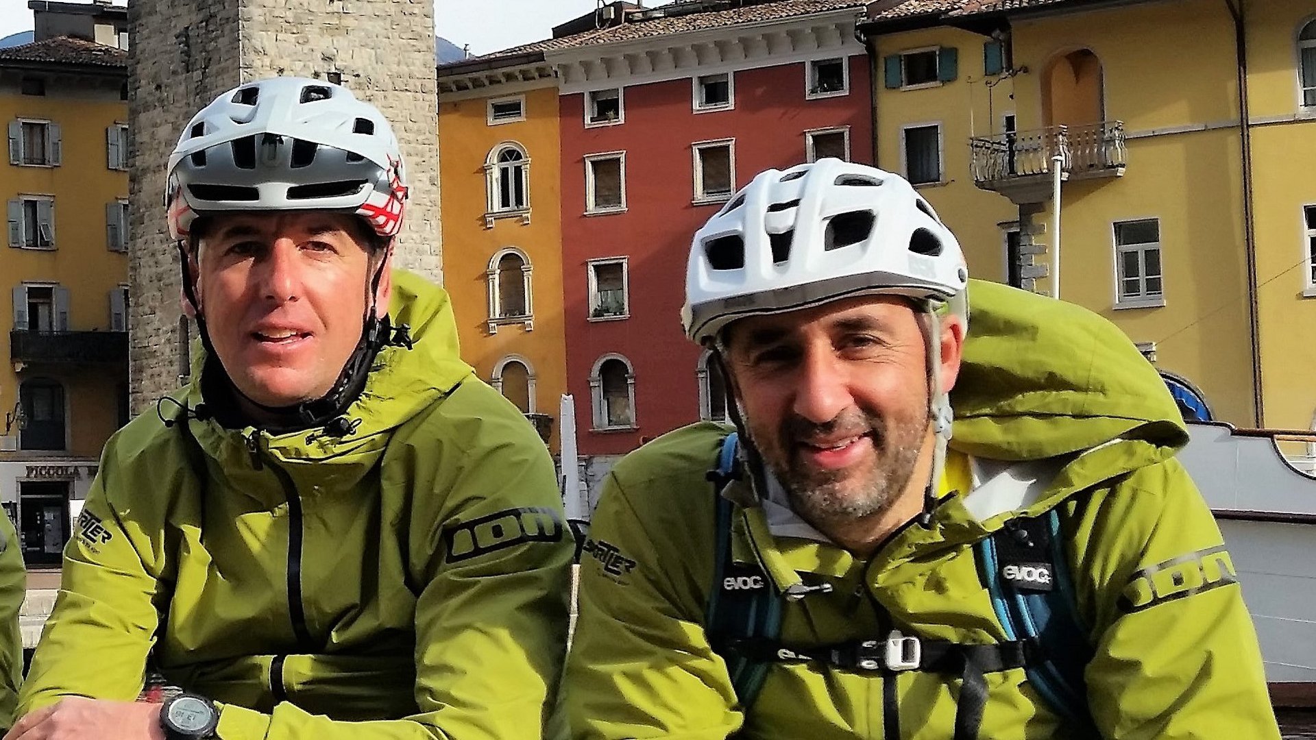 Zwei Radfahrer in grünen Jacken und weißen Helmen sitzen vor bunten Häusern