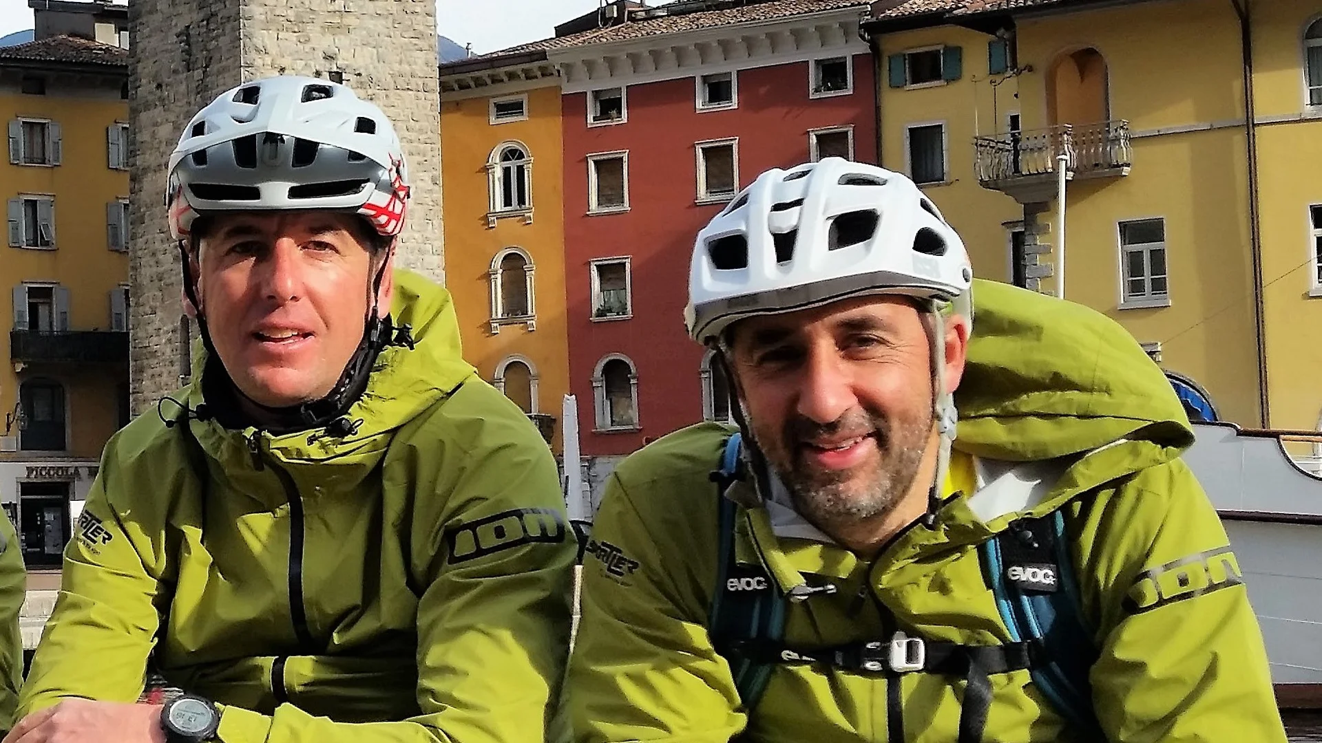 Zwei Radfahrer in grünen Jacken und weißen Helmen sitzen vor bunten Häusern