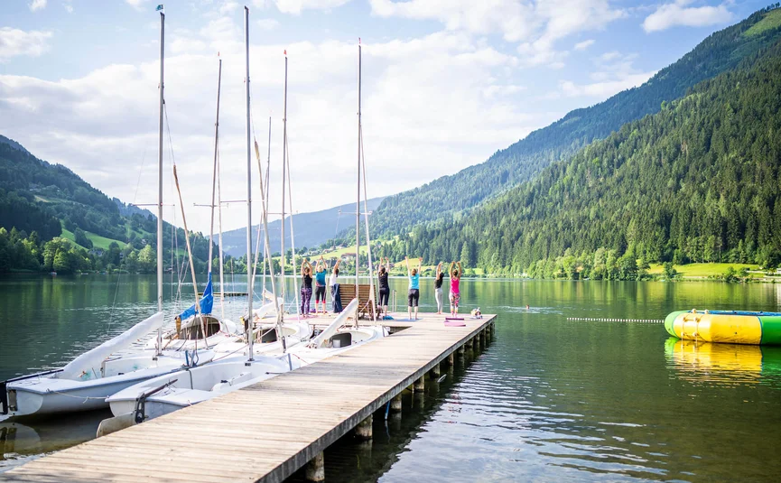 Menschen machen Yoga auf einem Steg neben Segelbooten an einem Bergsee