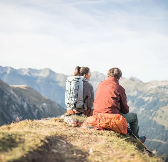 Zwei Wanderer sitzen mit Rucksäcken und schauen auf Bergpanorama
