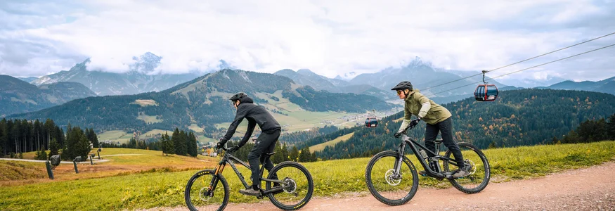 Zwei Radfahrer fahren auf einem Bergweg mit Seilbahn und Bergen im Hintergrund