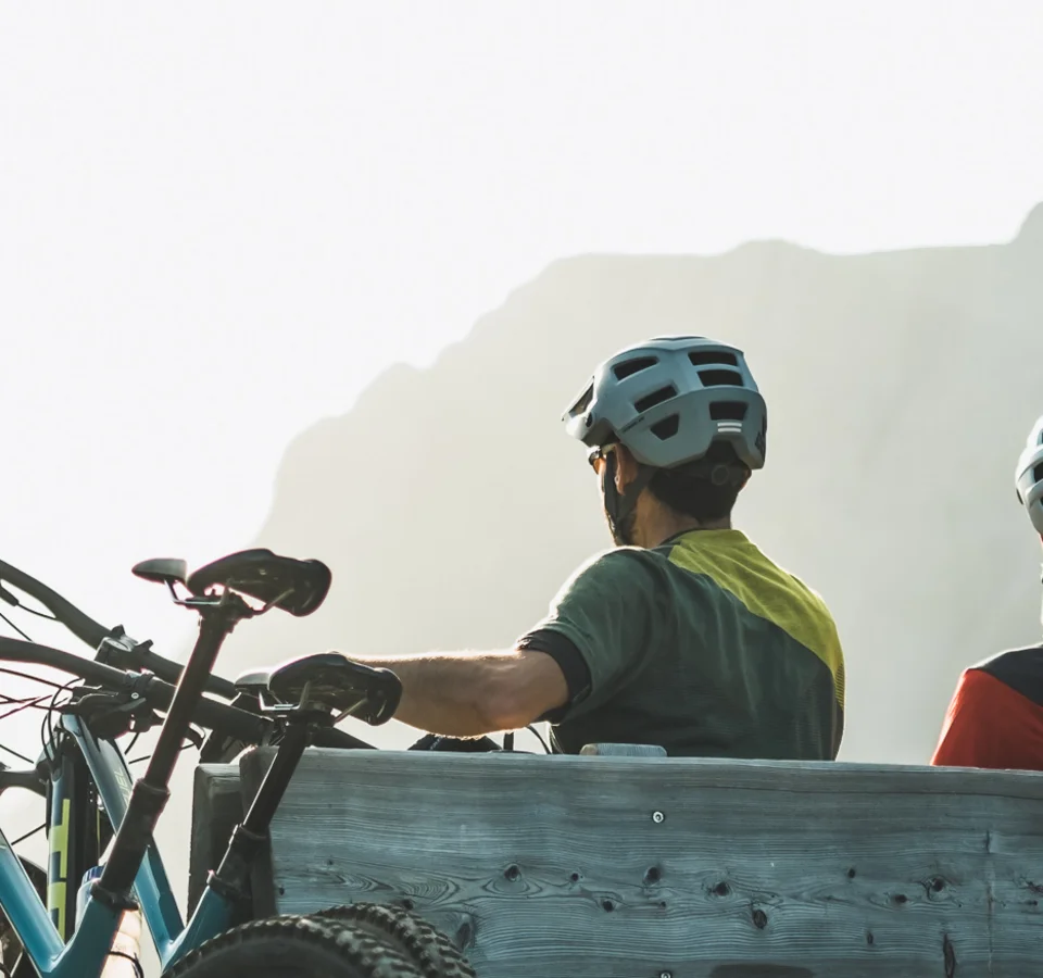 Zwei Radfahrer mit Helmen sitzen auf einer Holzbalkenbank vor Berglandschaft