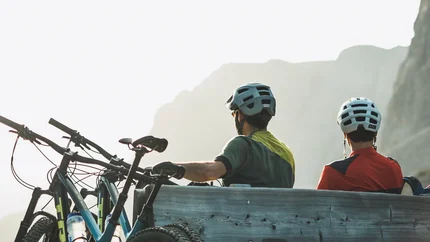 Zwei Radfahrer mit Helmen sitzen auf einer Holzbalkenbank vor Berglandschaft