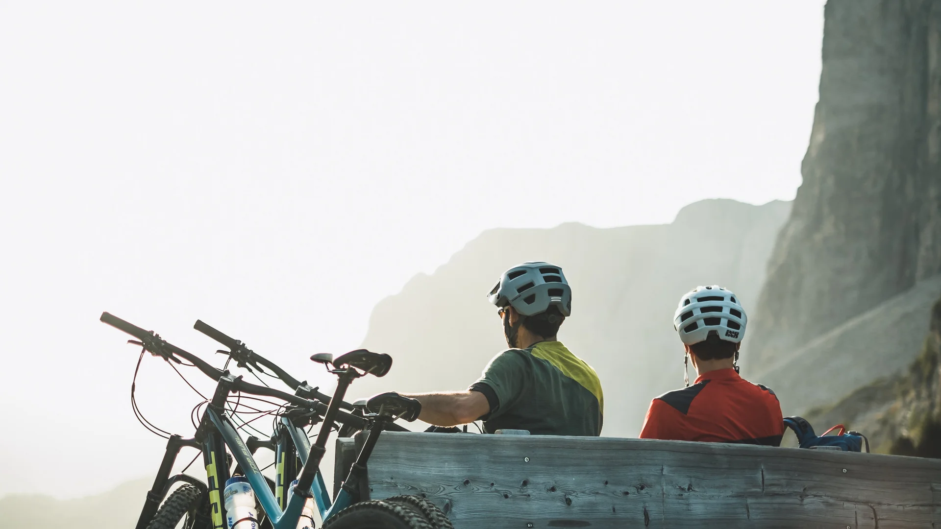 Zwei Radfahrer mit Helmen sitzen auf einer Holzbalkenbank vor Berglandschaft