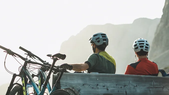 Zwei Radfahrer mit Helmen sitzen auf einer Holzbalkenbank vor Berglandschaft