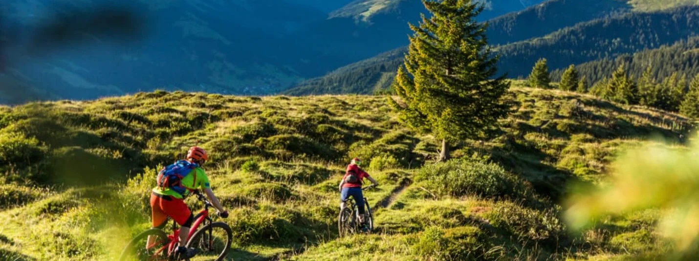 Two cyclists biking on a green mountain trail with mountains in the background