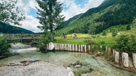 Mit Martina & Flo zum Mountainbiken im LifeSport Hotel Hechenmoos © Martina Wunderle / Flo Distl Zwei Radfahrer fahren auf einem Weg neben einem Bach und Bergen im Hintergrund