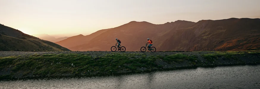 Die besten Touren & Trails der Alpen © Tobias Köhler Zwei Radfahrer fahren bei Sonnenuntergang an einem Bergsee vorbei
