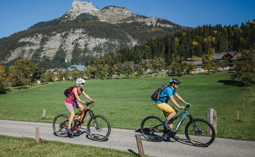 Two cyclists riding on a country road with mountain backdrop on a sunny day