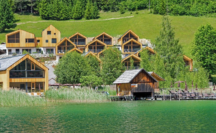 See-Aktivhotel Regitnig - Hotel & Chalets **** © Regitnig GmbH Holzferienhäuser am See mit grünen Bäumen und Hügeln im Hintergrund
