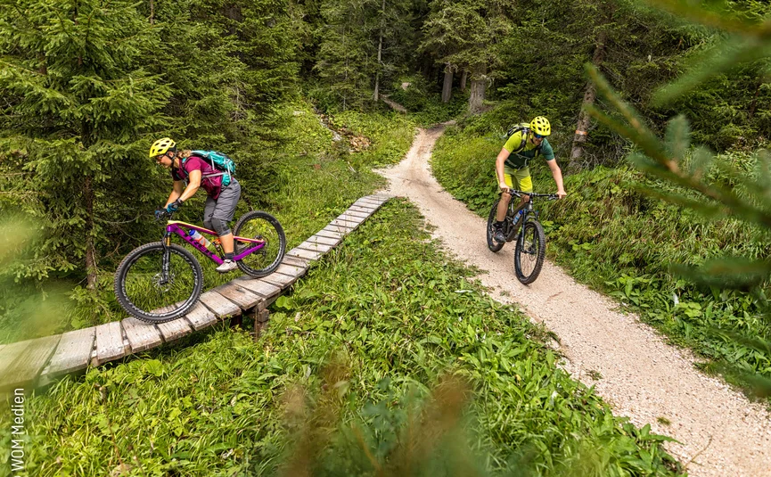 Zwei Mountainbiker fahren auf Waldweg, einer auf Holzbrücke, beide mit Helm und Rucksack