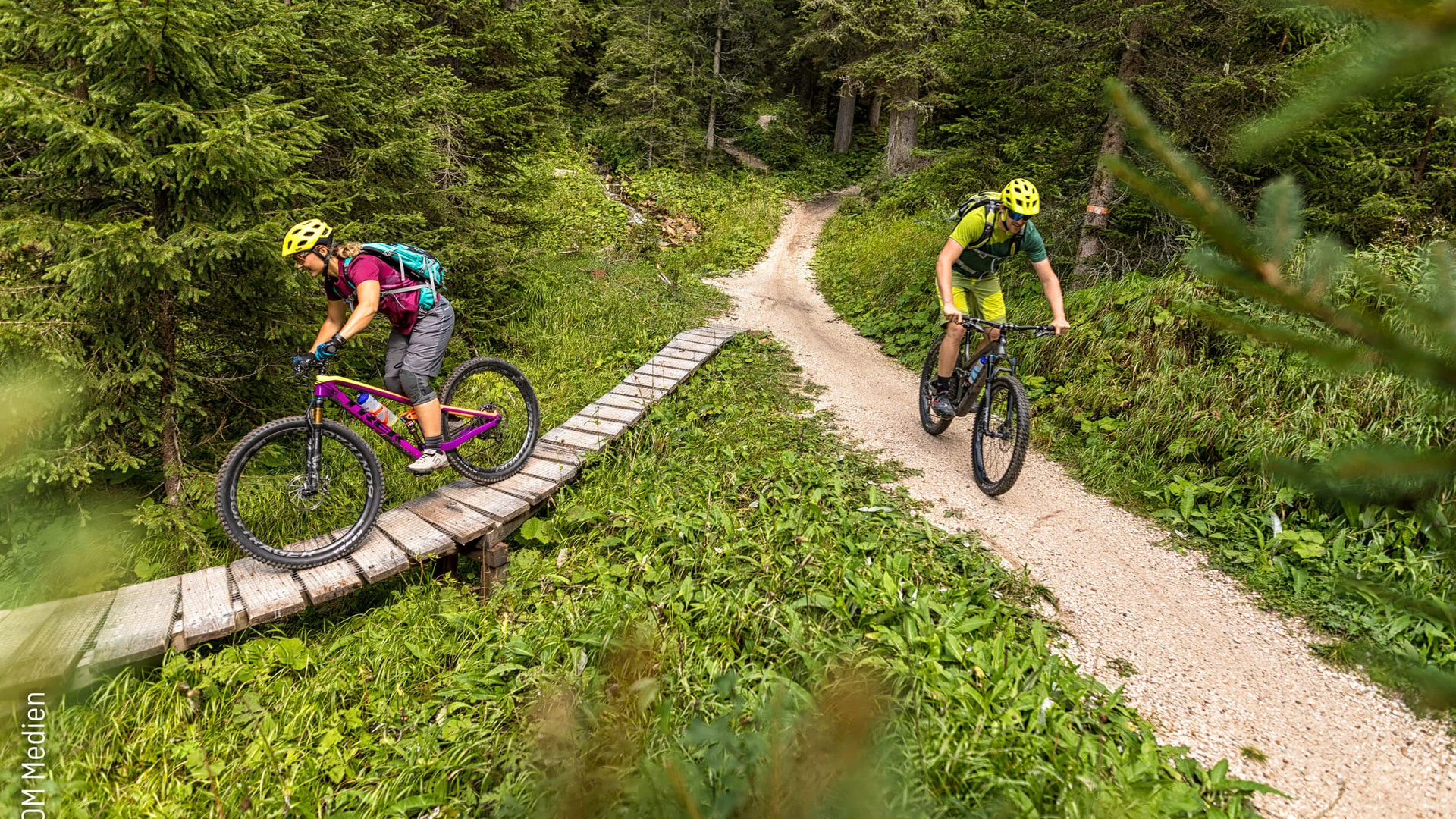 Two mountain bikers ride on forest trail, one crossing wooden bridge, both wearing helmets and backpacks
