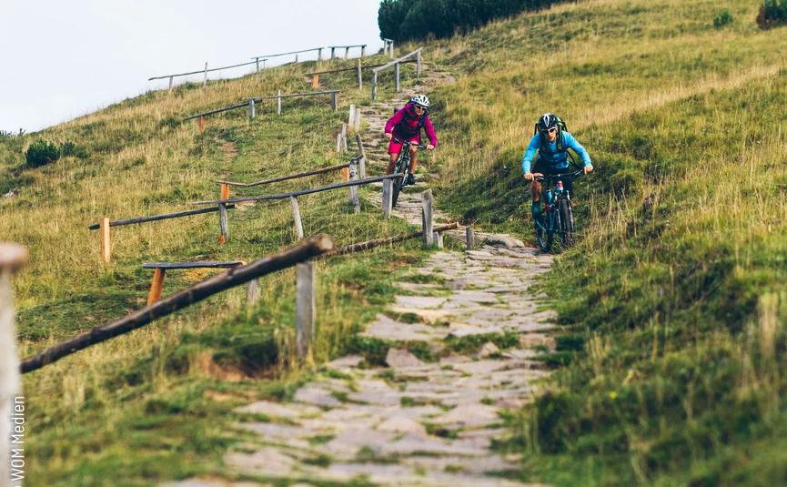 Two cyclists ride downhill on a rocky trail through green hills.