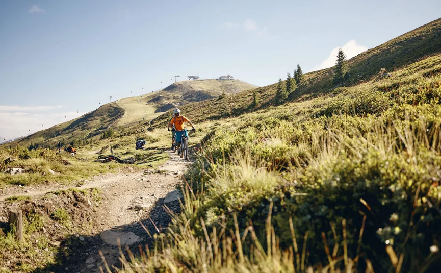 Cyclists riding on a sunny mountain trail with cable cars in the background