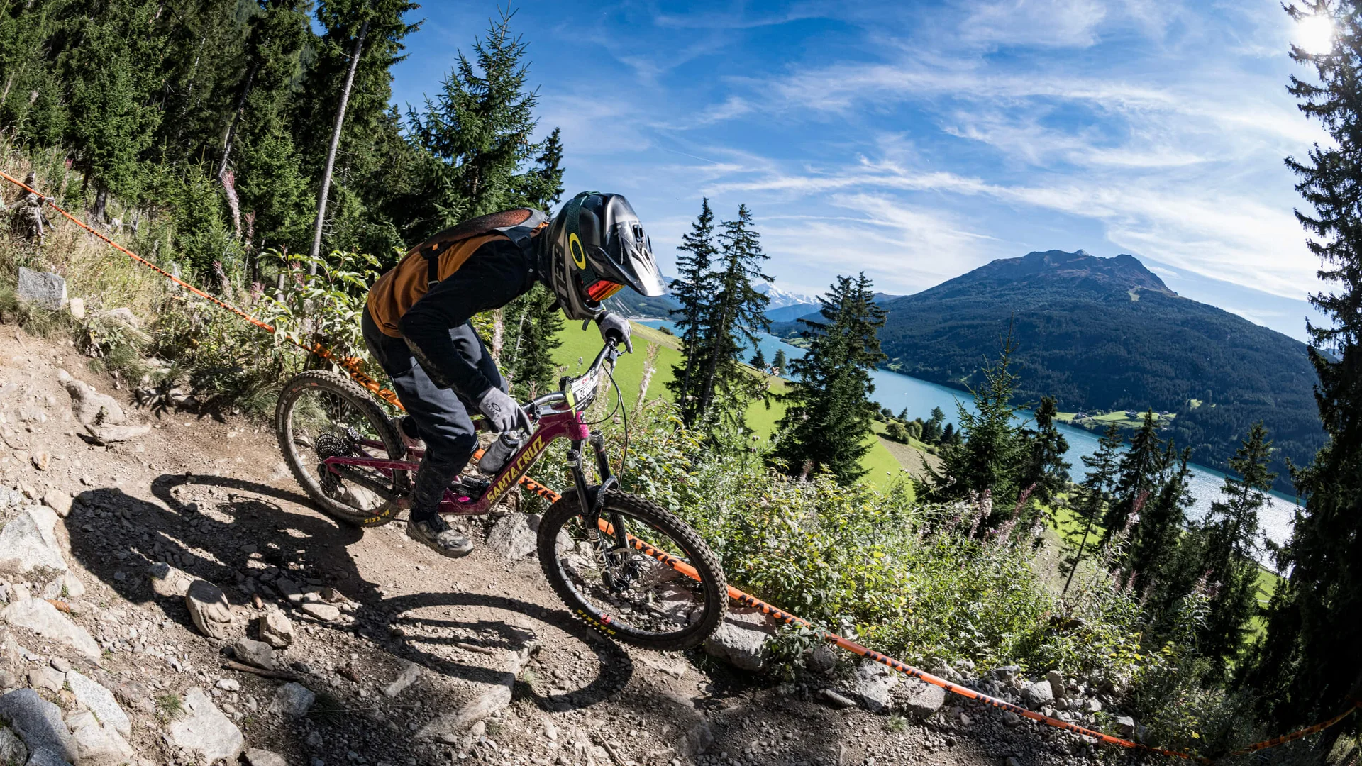 Mountainbiker fährt auf felsigem Waldweg mit Berg und See im Hintergrund