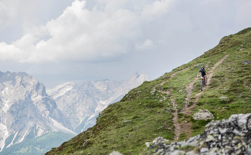 Mountainbiker fährt auf schmalem Pfad an Berghang mit Felsen und Wolken