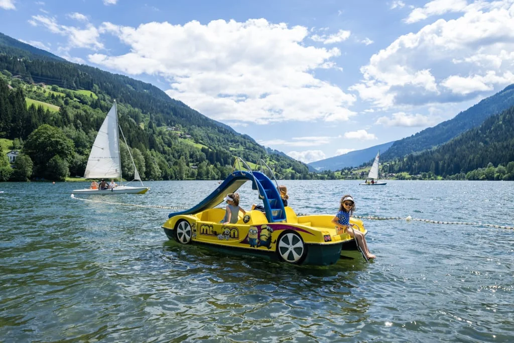 Brennseehof Happy Family Woche Kinder auf gelbem Tretboot mit Rutsche auf See mit Segelbooten und Bergen