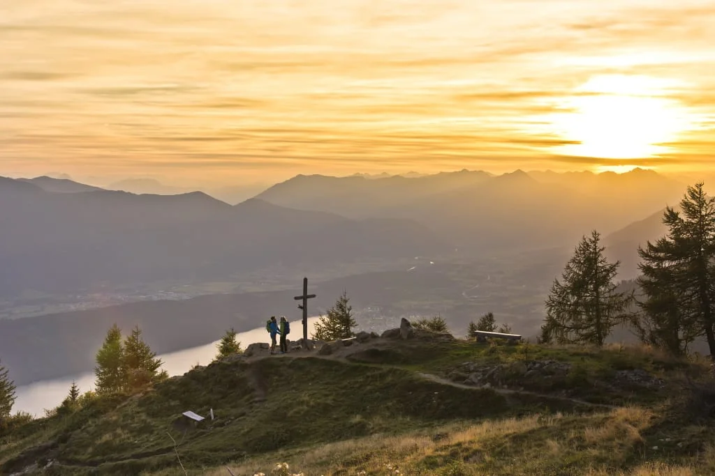 Zwei Wanderer neben Gipfelkreuz bei Sonnenuntergang in den Bergen